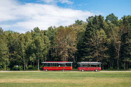 Chambord, Canada - August 18, 2025: Red tourist shuttle buses circulating through Val-Jalbert Historical Village amid pine and birch trees under blue skyのeditorial素材