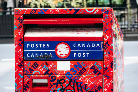 Montreal, Canada - August 14, 2025: A bright red Canada Post mailbox covered with colorful stickers, graffiti, and alphanumeric patterns, standing on a downtown sidewalkのeditorial素材