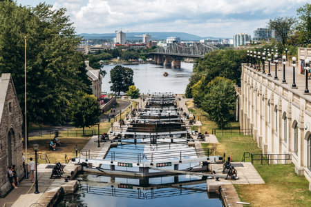 Ottawa, Canada - August 13, 2025: Wide view of the Rideau Canal locks with stone buildings, pathways, and people walking along the water, with the Ottawa Riverのeditorial素材
