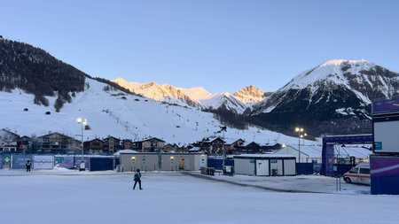 Livigno, Italy - February 8, 2026: Milano Cortina 2026 Olympic venue with cabins, barriers, person walking on snowy field amid Alps at sunsetのeditorial素材