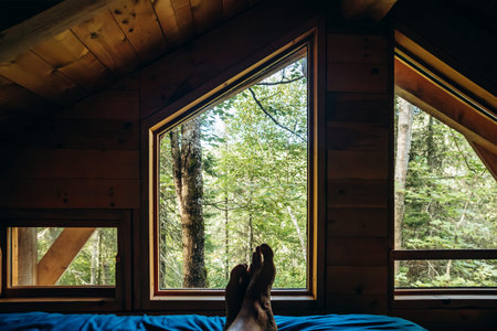 Male bare feet resting on a bed inside a wooden forest cabin, with a view of green trees through...