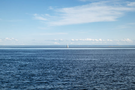 Wide view of a river meeting another body of water, with a solitary white sailboat on calm blue water under a clear summer skyの写真素材