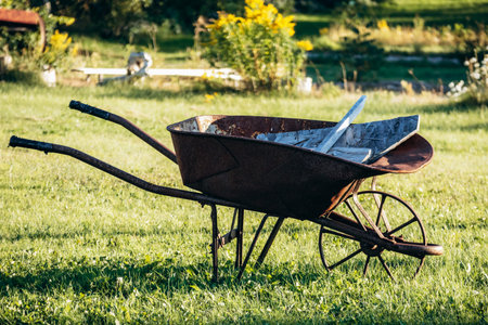 Rusty vintage metal wheelbarrow standing on green grass in a rural garden setting under warm summer sunlight, Baie-Sainte-Catherine, Canadaの写真素材