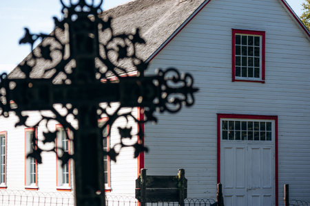 Ornate metal cross in soft focus in the foreground with historic white wooden church in focus behind it at the closed cemetery of Saint Firmin Historic Village in Quebecの写真素材