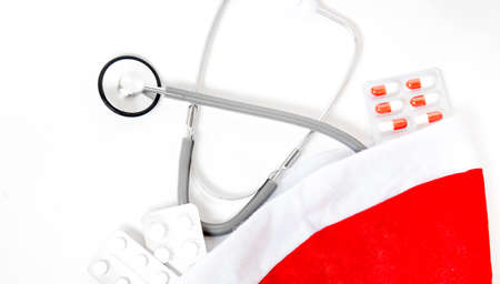 Medicine christmas new year concept banner during coronavirus covid-19 time. Top view of medical pills, disposable protective face mask, gift box, stethoscope in santa hat on red background.の写真素材