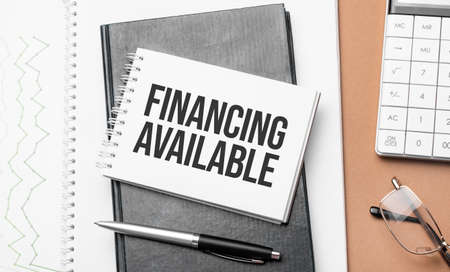 FINANCING AVAILABLE on notepad and various business papers on brown background. Brown glasses and magnifier with notepad.の写真素材