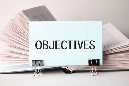 A white card with the text OBJECTIVES stands on a clip for papers on the table against the background of books. Defocusの写真素材