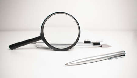 On a light wooden background, a magnifying glass, documents under the clips for papers and a pen.の写真素材