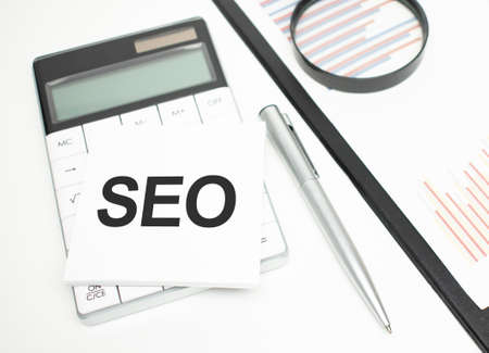 The word SEO is written in a daily diary between a calculator and a cup of coffee on a light wooden table. Business conceptの写真素材
