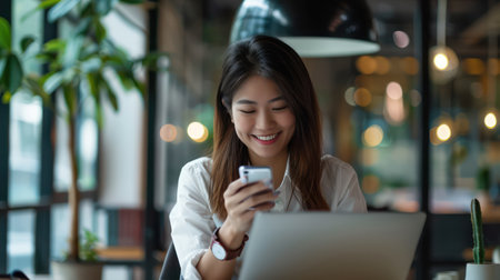 Young smiling asian businesswoman using and looking at mobile phone during working on laptop computer at modern officeの素材
