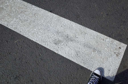 Male foot stepping over a wide white road marking, with the rubber foot tip of the shoe completing...