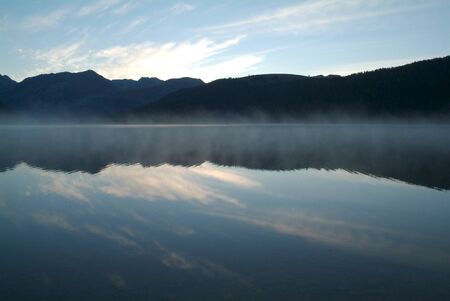 Symmetrical mountains reflected in the lake with beautiful blue skies and dense mistの写真素材