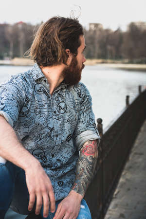 Portrait of young bearded handsome man with tattooed body looking sexually at camera near a lakeの写真素材