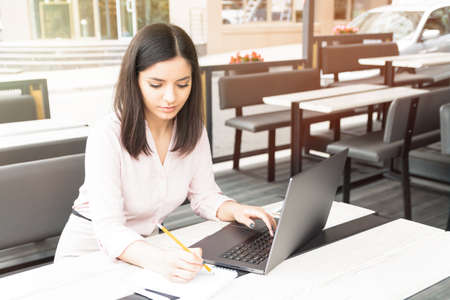 Young woman sitting in a cafe working on computer and writing in a notebook.の写真素材