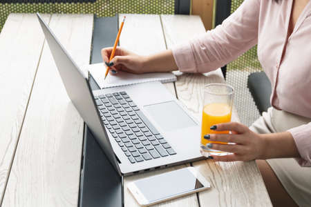Close up portrait of a young woman working on laptop and writing, holding a glass of orange juice.の写真素材