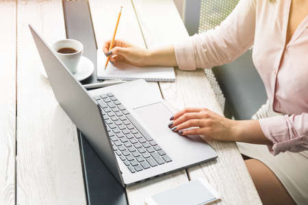 Business, education, people and technology concept - close up of female hands working on laptop, writing on paper, coffee on the tableの写真素材