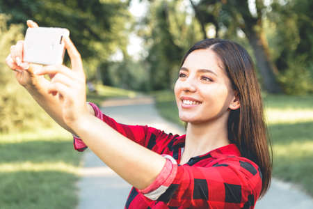 Portrait of a beautiful brunette girl in a red, plaid shirt, taking selfie, video chattingの写真素材