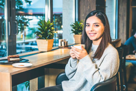 Young beautiful woman holding a cup of coffee or tea sitting by window. Cake on tableの写真素材