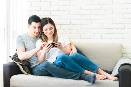 Relaxed couple using a generic mobile phone together sitting on a sofa in the living room at homeの写真素材