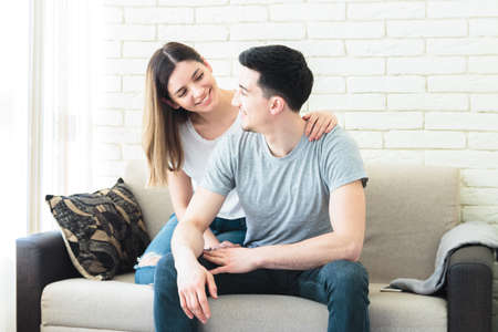 Cute couple in love hugging on the sofa indoors at home, wearing casual outfitsの写真素材