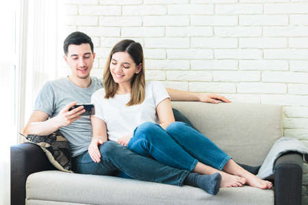 Relaxed couple using a generic mobile phone together sitting on a sofa in the living room at homeの写真素材
