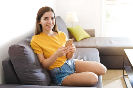 beautiful woman in a yellow t-shirt sitting on the sofa bed in a bright room, smiling and holding a phoneの写真素材
