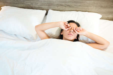 high angle view of a sleepy woman rubbing eyes while lying in bed at homeの写真素材