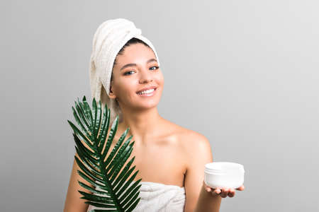 beauty woman with clean skin and towel holding white cream jar and palm leafの写真素材