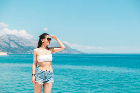 time to travel. beautiful woman in white topic and jeans shorts alone on seaside looking to the blue ocean and enjoying the viewの写真素材