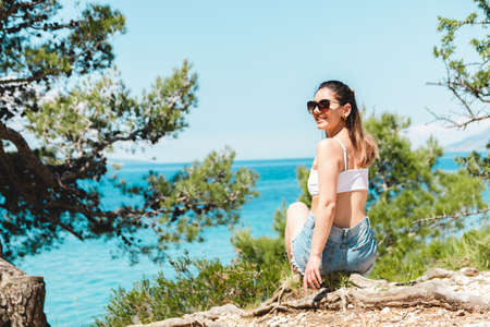 beautiful young woman traveler enjoying a sea view scene from a hill between pine treesの写真素材