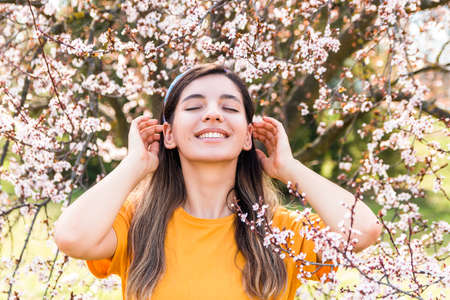 cheerful young woman with eyes closed enjoying beginning of spring against bloomy fruit tree with pink flowers in parkの写真素材