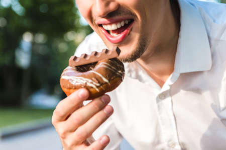 close up view of handsome caucasian young man in white shirt enjoying delicious chocolate donut in city park. junk but tasty food for positive mood.の写真素材