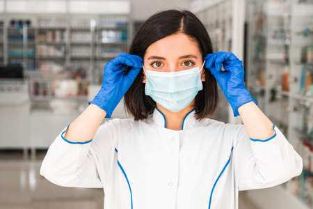 close portrait female pharmacist in pharmacy putting on face a protective mask getting to work to help peopleの写真素材