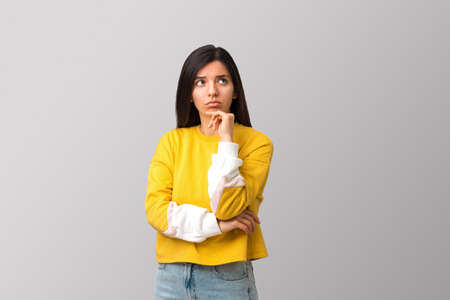 thoughtful young attractive multi ethnicity woman in trendy yellow sweater against light grey background holding her chin and looking upの写真素材