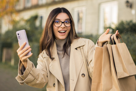 Portrait of attractive happily surprised woman with glasses holding eco-carton shopping bags and a phone in urban streetの写真素材