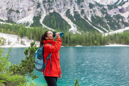 Hydration while on track. Attractive woman hiker in red raincoat drinking water near a lake and mountainsの写真素材
