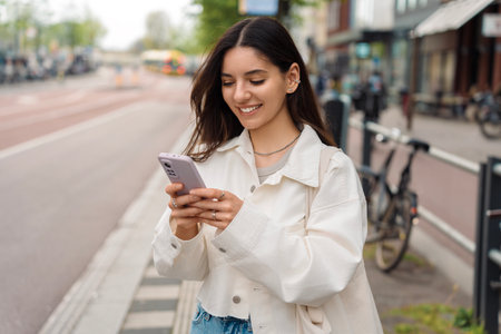 Everyday commute. Cheerful young woman student using the app on her phone while waiting for the public transportの写真素材