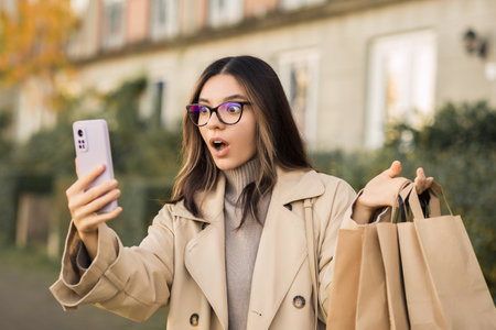 Shocking sales. Surprised fashionable woman checking her phone holding paper shopping bags in the streetの写真素材