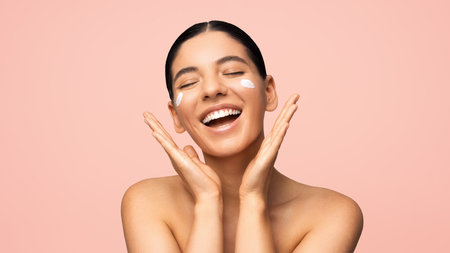 Face skincare. Beautiful multi-ethnic woman with cheerful smile applying moisturizer on face. Beauty portraitの写真素材