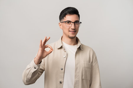 Sealed with confidence. A man in eyeglasses rocking a trendy beige shirt, flashing an OK gesture. Effortless style meets self-assured energyの写真素材