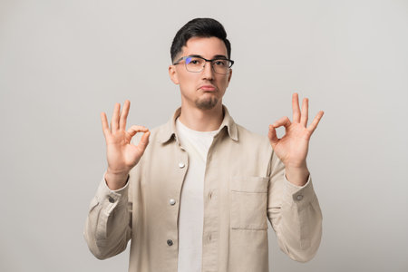 Unshakable confidence. A man in eyeglasses and a beige overshirt holding both hands in an OK gesture, exuding bold self-assurance. Modern style meets unstoppable charismaの写真素材
