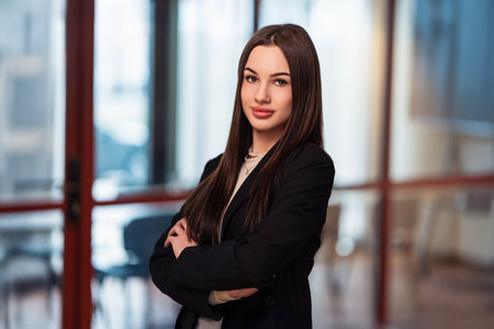 Next-gen leader vibes. A young businesswoman stands with arms crossed in a contemporary office, exuding confidence and ambition. Youthful drive meets corporate styleの写真素材