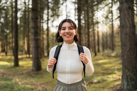 Hiking woman in a white top and black backpack smiling in a forest, blending outdoor adventure with wellness and carefree lifestyle vibesの写真素材