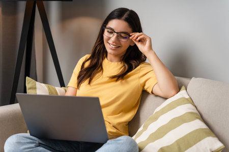 Woman sits comfortably on the couch in glasses, focused on her laptop as she works remotely, checks online orders, and scrolls through weekend dealsの写真素材