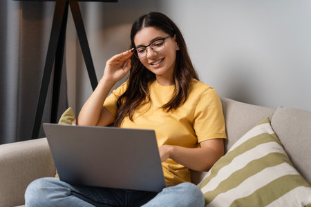 Relaxed on the sofa, a woman in eyeglasses switches between freelance tasks, online shopping tabs, and catching up on personal emailsの写真素材