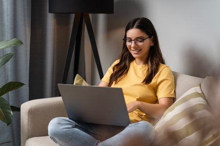 In soft weekend light, a woman on the couch multitasks between client work, casual internet surfing, and adding new finds to her online cartの写真素材
