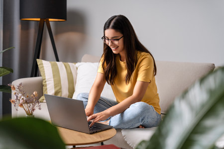 Young woman with glasses leans into her laptop, managing remote work deadlines while browsing for home items and checking in on her courseworkの写真素材