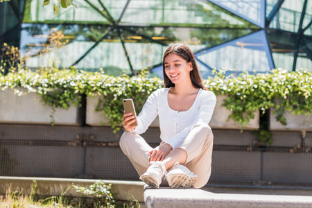 Relaxed young woman sitting outside with her phone, soaking in sunshine while managing wellness apps, chats, and peaceful productivity on the goの写真素材