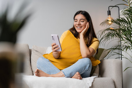 Digital self-care at home. Latina Indian woman in her 20s on sofa with phone, tuning into online therapy or study, blending mental wellness and digital learning in a chill home setupの写真素材