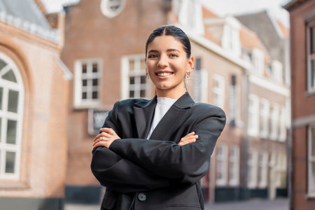 Confident young brunette realtor in sharp black suit posing with arms crossed outdoors, perfect for real estate marketing, career success, city professionalism, or personal brandingの写真素材